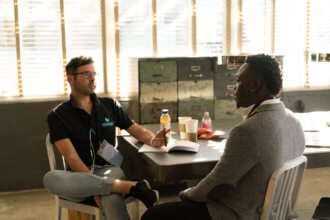 Two guys sitting across from each other, near a desk, having a conversation.