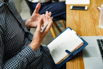 Guy sitting at a desk holding a pen and balancing a notebook in his lap, explaining something.