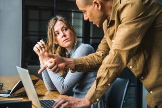 two people sitting at a desk conversing over something on the screen of the laptop in front of them.