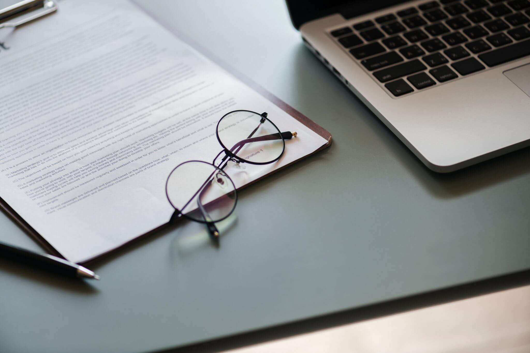 business document on clipboard next to a laptop and reading glasses.