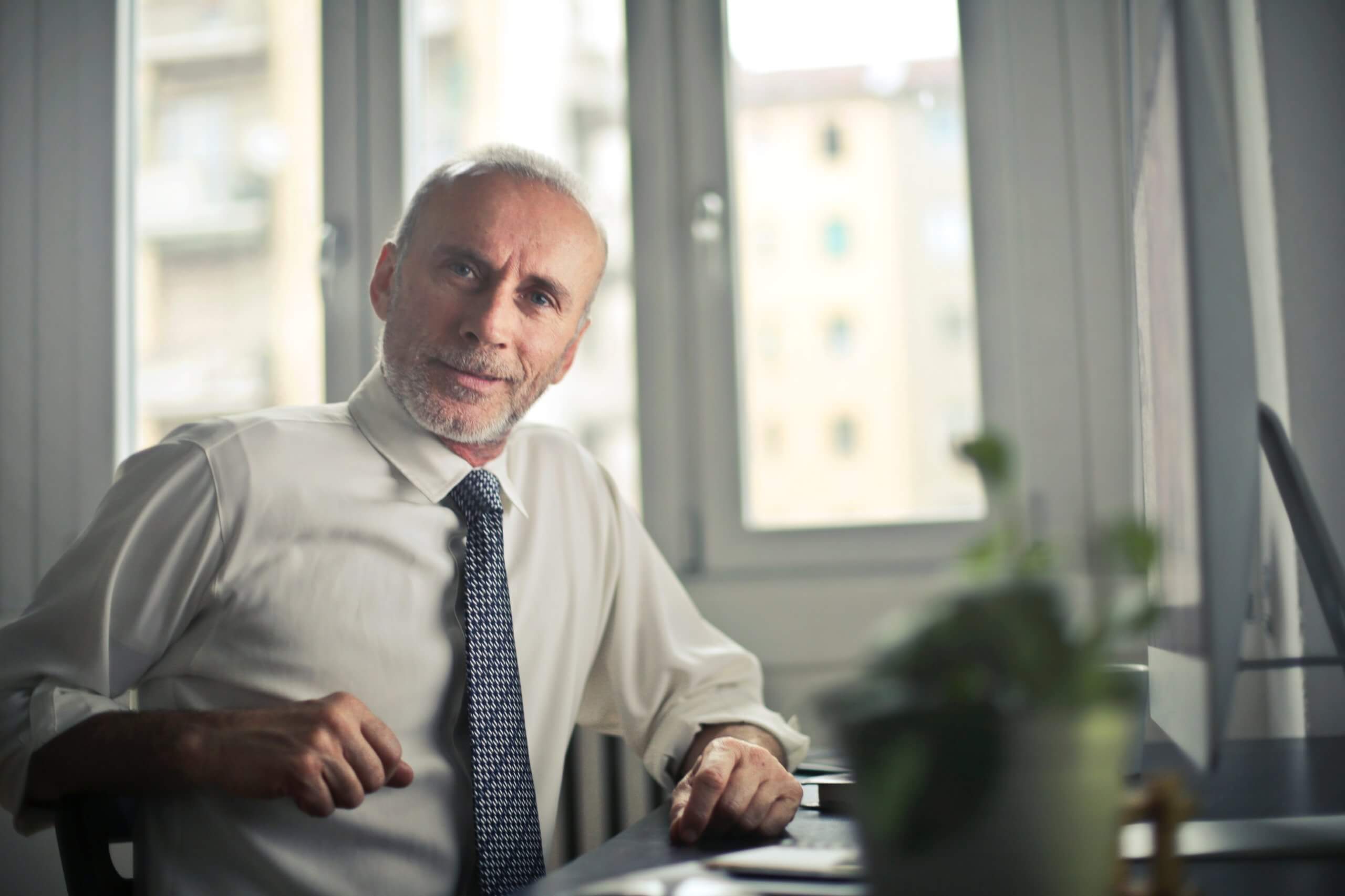 older gentleman in a business shirt and tie sitting at a desk looking at camera