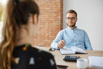 Business concept. Job interview. Adult bearded male hr manager in glasses and shirt sitting in light office in front of brunette caucasian woman, asking questions about previous work place