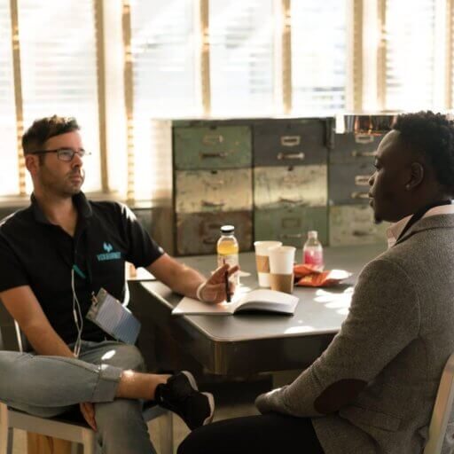 Two guys sitting across from each other, near a desk, having a conversation.