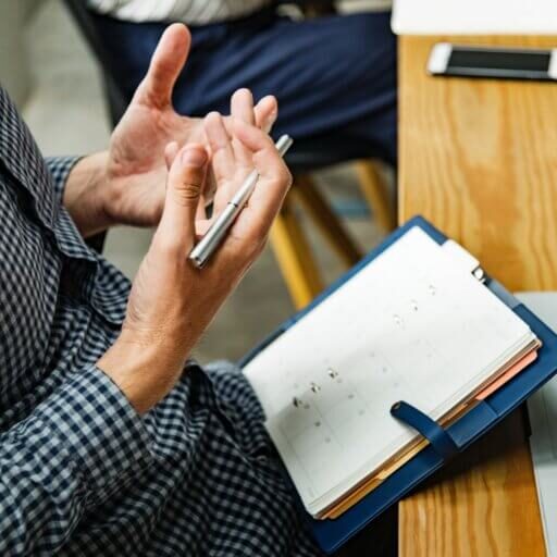 Guy sitting at a desk holding a pen and balancing a notebook in his lap, explaining something.