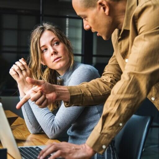 two people sitting at a desk conversing over something on the screen of the laptop in front of them.