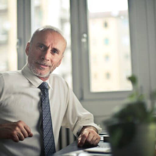 older gentleman in a business shirt and tie sitting at a desk looking at camera